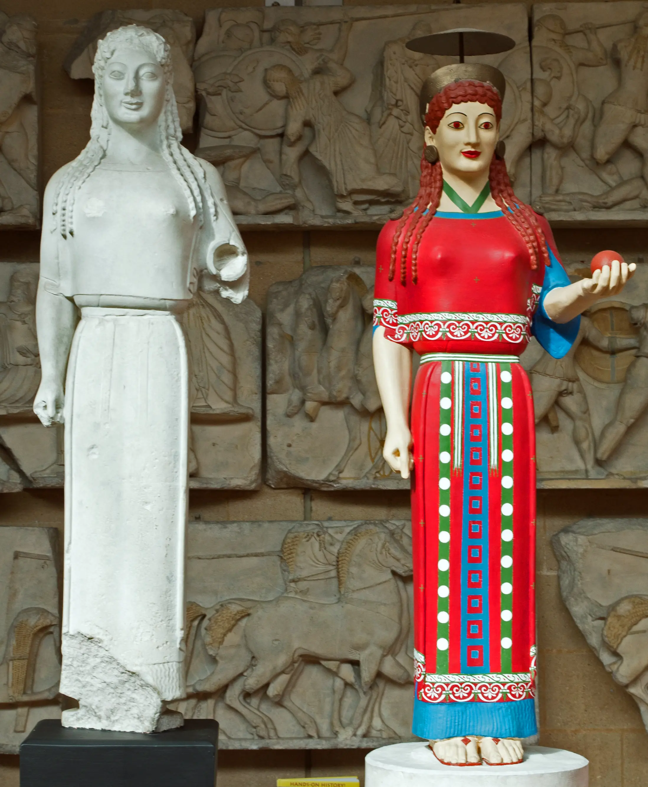 Two museum casts of the ancient Greek Peplos Kore shown side by side: one pale plaster cast and one reconstruction with painted hair, skin, clothing, and patterned decoration.